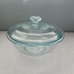 May include: A clear glass bowl with a matching lid. The bowl is round with a slightly flared rim. The lid has a small handle and a textured design. The glass has a slight blue-green tint.