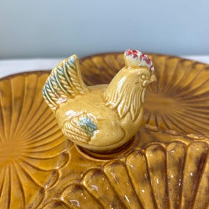 May include: A ceramic serving dish with a decorative chicken figurine. The dish is a warm yellow-brown color with a textured, fan-like pattern. The chicken has a red comb and blue accents.