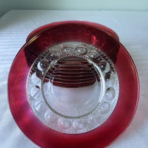 May include: A stack of round glass plates featuring a deep red translucent border and a clear center. The clear portion of the plates is decorated with a repeating pattern of raised circular dots and a textured rim detail around the inner edge.