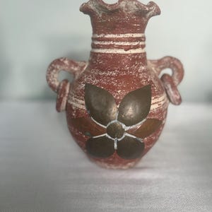 May include: A small, terracotta-colored vase with a textured surface and two handles. The vase features a decorative metal flower design on the front. The rim and handles have a distressed, aged appearance, adding to its rustic charm.