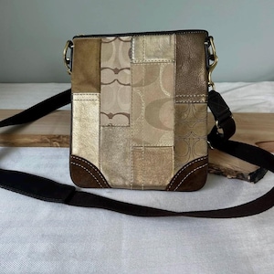 May include: A square crossbody bag featuring a patchwork design of metallic gold leather, tan suede, and beige fabric with a repeating logo pattern. The bag includes dark brown suede corner accents, a top zipper closure, and a long adjustable black strap.