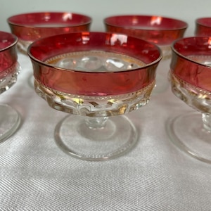 May include: Set of vintage clear glass champagne coupe glasses with a ruby red rim and gold detailing. The glasses have a decorative base and are arranged on a white surface. Perfect for serving champagne or cocktails.
