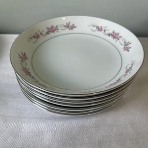 May include: A stack of white ceramic bowls with a floral design. Each bowl has a silver rim and a delicate pattern of pink flowers and gray accents around the edge. The bowls are stacked on a white surface.