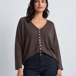 May include: A dark brown long-sleeve jumper featuring a V-neckline and a vertical row of small, light-coloured buttons running down the centre front. The jumper has a relaxed fit with dropped shoulders and is paired with dark blue denim jeans.