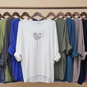 Op de afbeelding: Een collectie shirts met lange mouwen en vleermuismouwen hangend aan een houten rek. De shirts zijn verkrijgbaar in diverse effen kleuren zoals zwart, wit, blauw, groen, grijs en magenta. Het witte shirt heeft een zilverkleurige hartafbeelding.