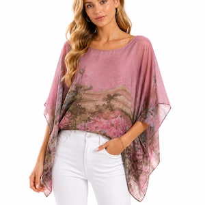 May include: A sheer, loose-fitting poncho-style top in a dusty rose hue, featuring a wide scoop neckline and short, flowing sleeves. The fabric displays a printed landscape scene with muted green and brown tones, paired with solid white jeans.
