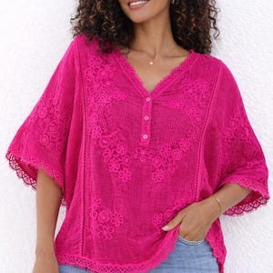 May include: A vibrant fuchsia-colored blouse with a V-neck and short sleeves. The blouse features intricate floral embroidery and lace trim along the neckline and hem. It has a button-down front and a relaxed fit, perfect for casual wear.