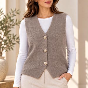 Puede incluir: Un chaleco sin mangas de color topo con un acabado de punto texturizado y difuso, con cuello en V y tres botones redondos de metal dorado en la parte delantera. El chaleco se lleva sobre una camiseta blanca de manga larga y pantalones beige claro.