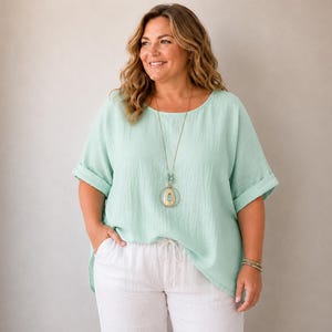 Peut inclure: Un haut vert menthe clair à manches courtes avec une encolure ronde et des manches à revers, associé à un pantalon blanc à cordon de serrage. Un long collier doré avec un pendentif circulaire et un bracelet de perles complètent l'ensemble.