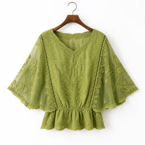 May include: A lime green blouse with a V-neck and short, flared sleeves. The blouse features intricate floral embroidery and a gathered waist. The fabric appears lightweight and semi-sheer, with a delicate lace trim along the neckline and sleeves.