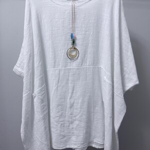 May include: A white, loose-fitting tunic with a round neckline, short sleeves, and two front pockets. A long necklace hangs over the centre, featuring a thin cord, small blue and green beads, and a large circular pendant with a metallic ring.