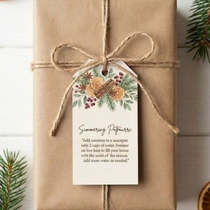 以下が含まれることがあります： 茶色の紙で包まれたギフトで、麻ひもで結ばれ、装飾的なタグが付いています。 タグには、乾燥オレンジ、シナモンスティック、緑の植物のテキストとイラストが描かれています。 タグには「Simmering Potpourri」と書かれています。