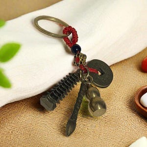 May include: A bronze-colored keychain with a key ring, red cord, and various charms. Charms include a pagoda, a brush, a coin, and a gourd. The keychain is designed with a traditional Asian aesthetic.
