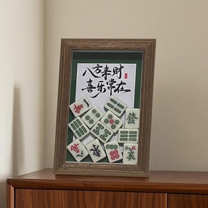 Può includere: Un'opera d'arte incorniciata con una collezione di tessere di Mahjong disposte davanti a uno sfondo verde. La cornice è di colore legno scuro. Le tessere sono bianche con simboli rossi, verdi e neri. Caratteri cinesi sono scritti sull'opera d'arte.