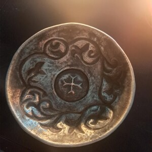 May include: A small, round, silver-toned decorative bowl. The bowl features an embossed design of swirling patterns and a central emblem. The rim has a slightly raised edge. The bowl's surface has a textured appearance.