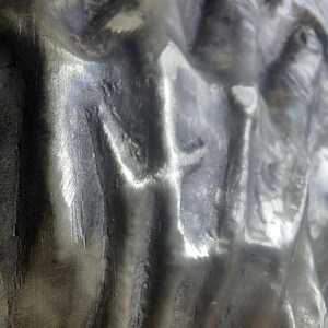 May include: Close-up of a metallic sculpture with a rough, textured surface. The metal has a silver sheen with areas of darker shading, creating depth and highlighting the sculpted forms. The image captures the details of the metalwork.