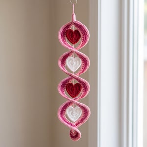 May include: A vertical hanging decoration featuring a crocheted spiral in shades of pink and magenta. The spiral structure contains four alternating crocheted hearts, two in solid red and two in solid white, suspended from a small metal ring.