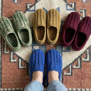 Può includere: Pantofole fatte a mano all'uncinetto in vari colori, tra cui verde, giallo senape, bordeaux e blu, disposte su un tappeto fantasia. Le pantofole presentano un design a coste. L'immagine mostra i piedi di una persona con pantofole blu.