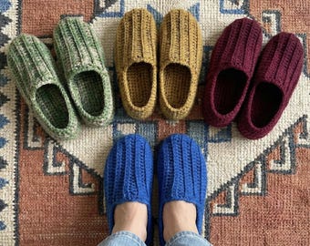 Puede incluir: Zapatillas de ganchillo hechas a mano en varios colores, incluyendo verde, mostaza, burdeos y azul, dispuestas sobre una alfombra estampada. Las zapatillas presentan un diseño acanalado. La imagen muestra los pies de una persona con zapatillas azules.