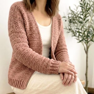 May include: A dusty rose crochet cardigan with long sleeves. The open-front sweater is made with a thick yarn, creating a textured appearance. The cardigan is worn over a white top and a cream-colored skirt.