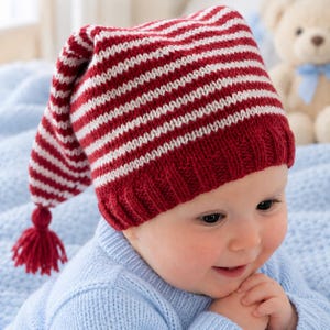 May include: A knitted baby hat featuring horizontal red and white stripes with a ribbed brim and a pointed crown topped with a small red yarn tassel. The hat is constructed with a soft knit texture and displays a classic stocking cap silhouette.