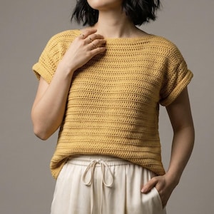 May include: Mustard yellow crochet top with short sleeves and a boat neck. The top is paired with off-white drawstring trousers. The crochet stitch creates a textured, horizontal striped pattern. The sleeves are cuffed. The overall style is casual and comfortable.
