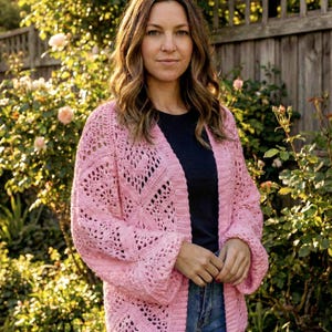May include: A pale pink crochet cardigan with an open front and intricate openwork pattern. The long-sleeved garment is worn over a dark top and blue jeans. The background includes a wooden fence and green foliage. The cardigan's design is visible.