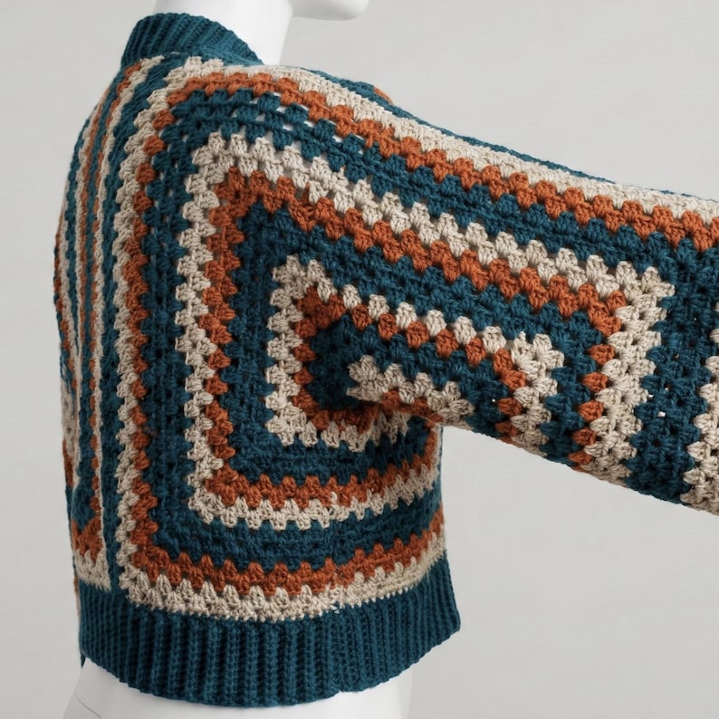 Pu&ograve; includere: Un maglione all'uncinetto color verde acqua, beige e ruggine. Il maglione presenta un motivo quadrato con maniche lunghe e orlo e polsini a coste. L'indumento &egrave; esposto su un manichino.