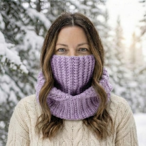 May include: A lilac-colored knitted cowl scarf worn around the neck and covering the lower face. The scarf is made of thick yarn with a ribbed texture. The person is wearing a cream-colored sweater. The background is a snowy forest.