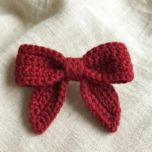 Puede incluir: Un lazo rojo tejido a mano. El lazo es de un color rojo intenso y rico y presenta una forma de lazo clásica con dos bucles y dos colas. Las puntadas de ganchillo son visibles, lo que le da al lazo una apariencia texturizada. Probablemente se utiliza como accesorio para el cabello o artículo decorativo.