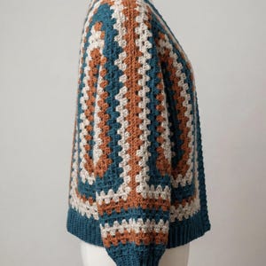 Pu&ograve; includere: Un cardigan all'uncinetto fatto a mano con un motivo a granny square. Il maglione presenta un design a blocchi di colore con quadrati verde acqua, beige e marroni. Le maniche e lo scollo sono verde acqua. Il cardigan &egrave; esposto su un manichino.