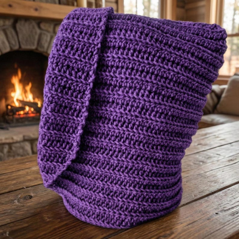 May include: A folded, handmade, purple crochet blanket. The blanket is made with thick yarn, creating a textured surface. The blanket is sitting on a wooden table. A fireplace with a burning fire is in the background.