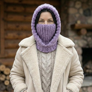 May include: A woman wearing a lavender knitted balaclava with a ribbed design, covering the head and neck. She is also wearing a cream-colored shearling coat and a cable-knit sweater. The background features a wooden structure.