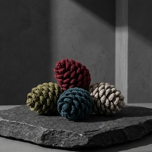 May include: Five handmade, decorative pine cones in shades of red, green, blue, and beige, arranged on a dark gray stone surface. The cones are made of a textured, woven material, and are a unique home decor item.