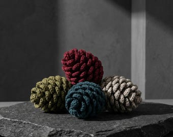 May include: Five handmade, decorative pine cones in shades of red, green, blue, and beige, arranged on a dark gray stone surface. The cones are made of a textured, woven material, and are a unique home decor item.