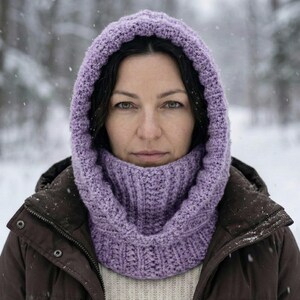 May include: A lilac-colored, knitted hood and cowl set. The hood frames the face, and the cowl wraps around the neck. The set is made of a thick, textured yarn, providing warmth. The background is a snowy forest.