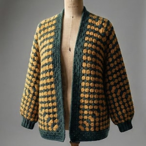 Puede incluir: Un cárdigan hecho a mano, abierto por delante, con un punto texturizado único. El suéter presenta un patrón de amarillo dorado y verde azulado oscuro, con el verde delineando los bordes y los puños. Las mangas son largas y el cárdigan se exhibe en un maniquí.