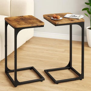 May include: Two side tables with a rustic brown tabletop and black metal frame. One table has a plate with two pastries and a magazine. The tables are designed to slide under a sofa.