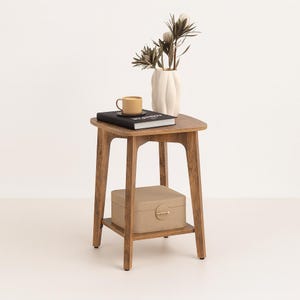 May include: A wooden side table with a lower shelf, holding a beige box with a gold clasp. The tabletop displays a yellow mug, a black book, and a white vase with dried floral stems. The table has a natural wood finish.