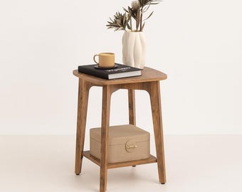 May include: A wooden side table with a lower shelf, holding a beige box with a gold clasp. The tabletop displays a yellow mug, a black book, and a white vase with dried floral stems. The table has a natural wood finish.