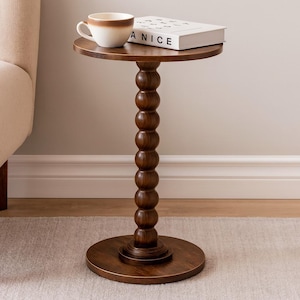 May include: A dark brown wooden side table with a round top and base, connected by a series of rounded wooden beads. A white and brown coffee cup and a book with the word "ANICE" are on the table.