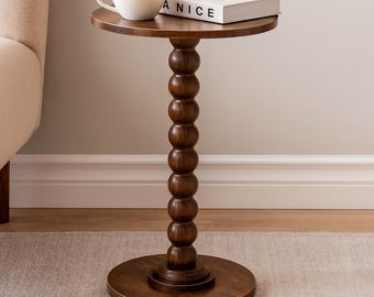 May include: A round dark brown wooden side table with a central pedestal featuring a stacked ball design. A white ceramic mug and a white book with the text A NICE printed on the cover are placed on the flat circular tabletop surface.