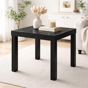 May include: A square, black side table with four sturdy legs. The table is topped with a white vase holding dried flowers, two books, and a candle in a glass holder. The table is approximately 24 inches square.