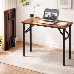 May include: A brown folding desk with black metal legs is set up with a laptop, a coffee cup, and a small notepad. Another desk is folded and standing upright in the background. The desk is on a light-colored rug.