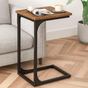 May include: A brown and black C-shaped side table with a rectangular tabletop. The tabletop has a wood grain finish. A cup of coffee and a magazine rest on the table. The table is next to a gray sofa.