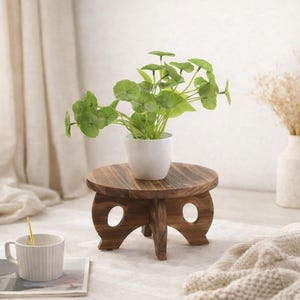 May include: A small, round wooden plant stand with a dark brown finish. A white ceramic pot with a green plant sits on top. The stand has three legs and two decorative cutouts. A white mug and a gold spoon are in the foreground.