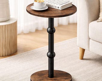May include: A round side table with a dark wood grain top and base, connected by a central black metal pedestal featuring two spherical accents. A white ceramic mug on a saucer and a white book are placed on the table surface.