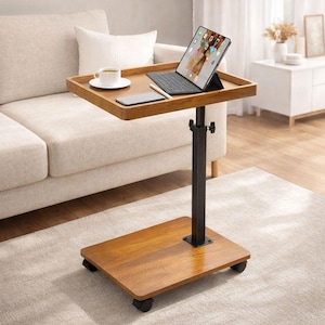 May include: A brown wooden laptop table with a black adjustable stand and rolling wheels. The table holds a tablet with a keyboard, a smartphone, a cup of coffee, and a pen. The table is next to a beige sofa.