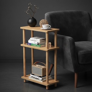 May include: A light wood side table with three shelves, holding books, a small radio, and decorative items. A black vase with branches, a woven ball, and a white teacup on a saucer are on the top shelf. A gray armchair is in the background.
