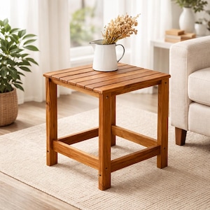 May include: A square wooden side table with a slatted top and four sturdy legs. The table is made of light brown wood and is holding a white pitcher with dried flowers. The table is 18 inches tall and 18 inches wide.